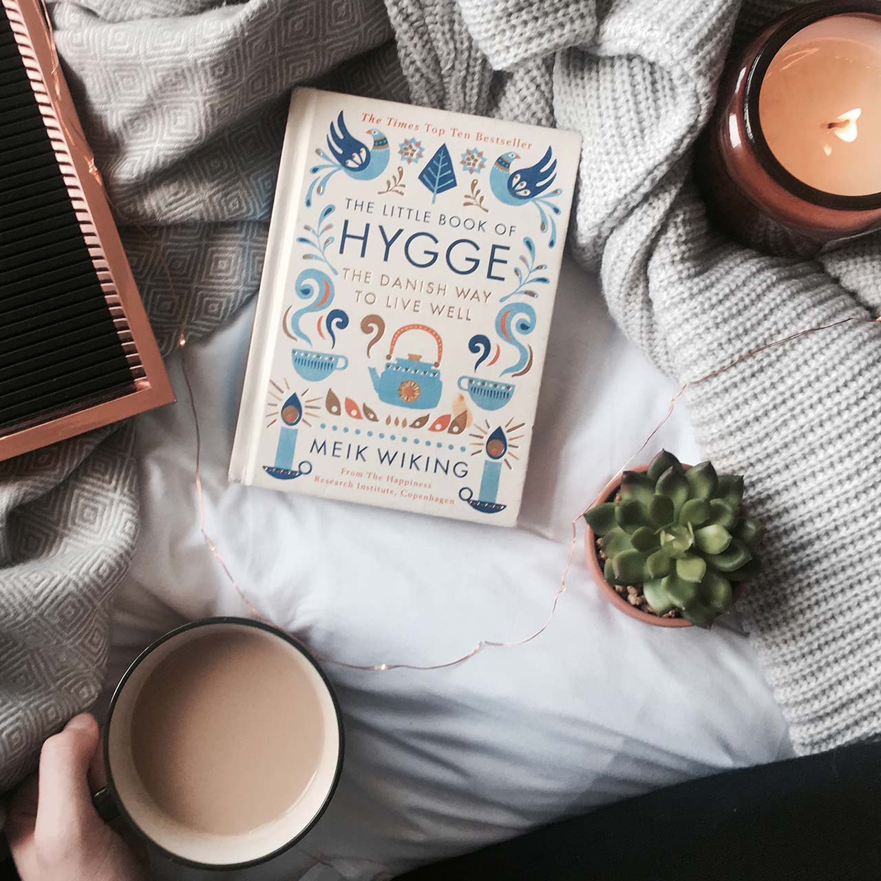 the little book of hygge by meik wiking on a bed surrounded by a cup of coffee, a burning candle and cosy blankets