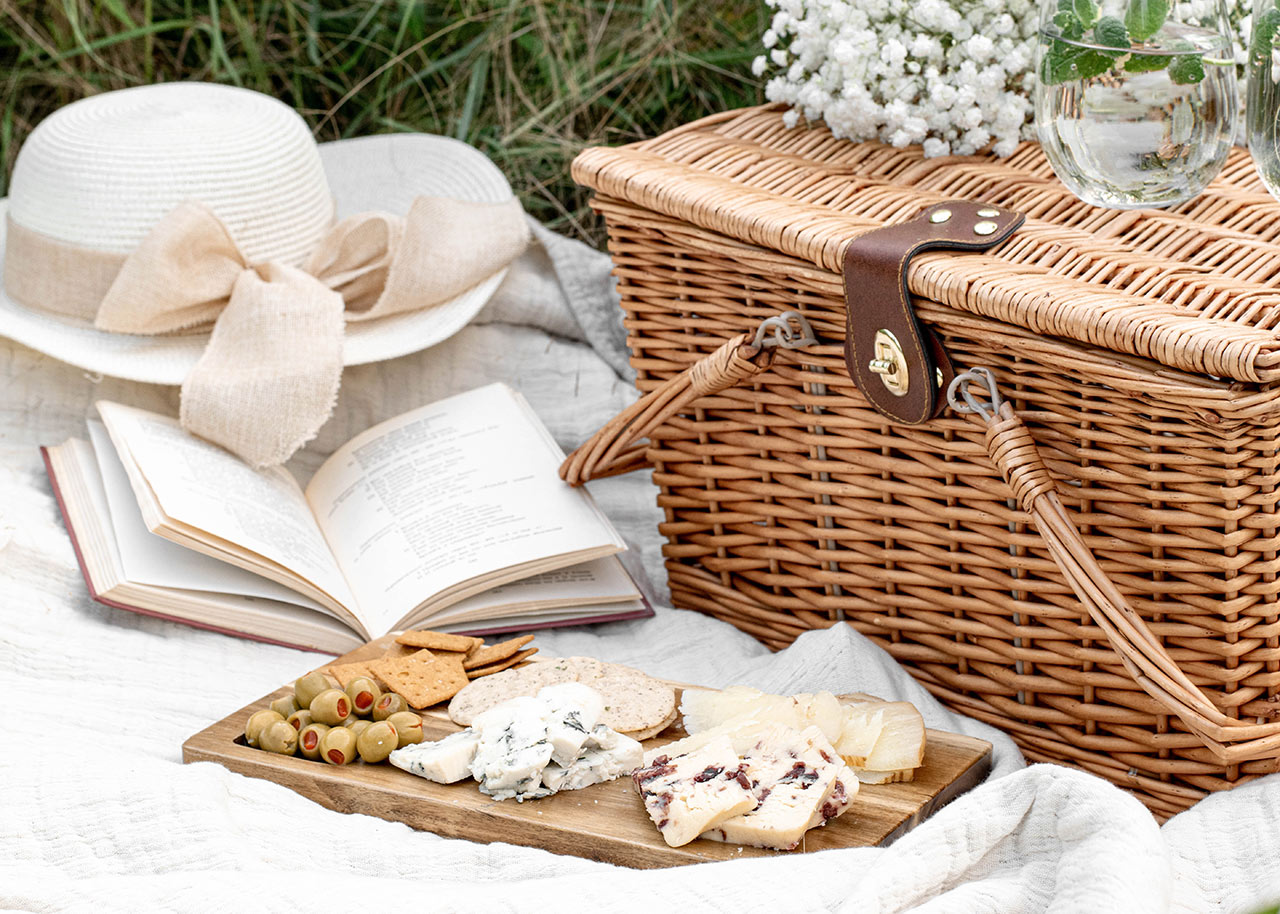 a blanket with a picnic hamper, food, flowers, a book and a sun hat