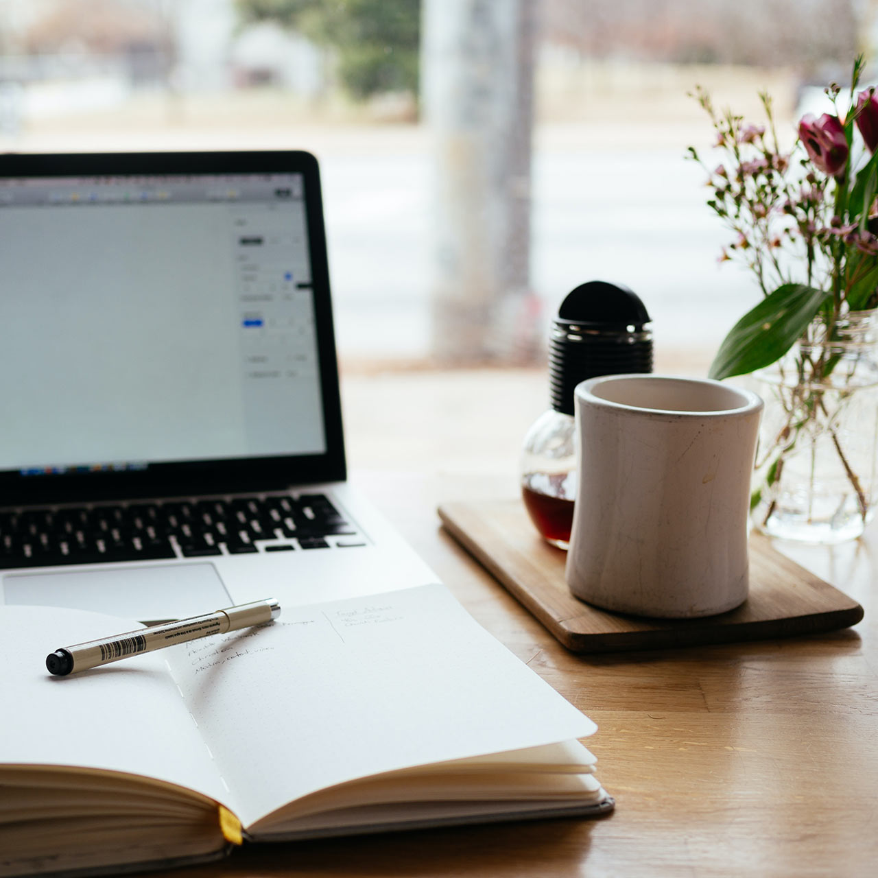 a cosy work set up with a laptop, a notebook and pen, coffee and a flower vase