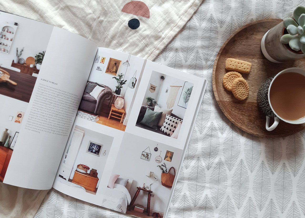 an open magazine on a bed with a small wooden tray with a cup of tea, cookies and a plant