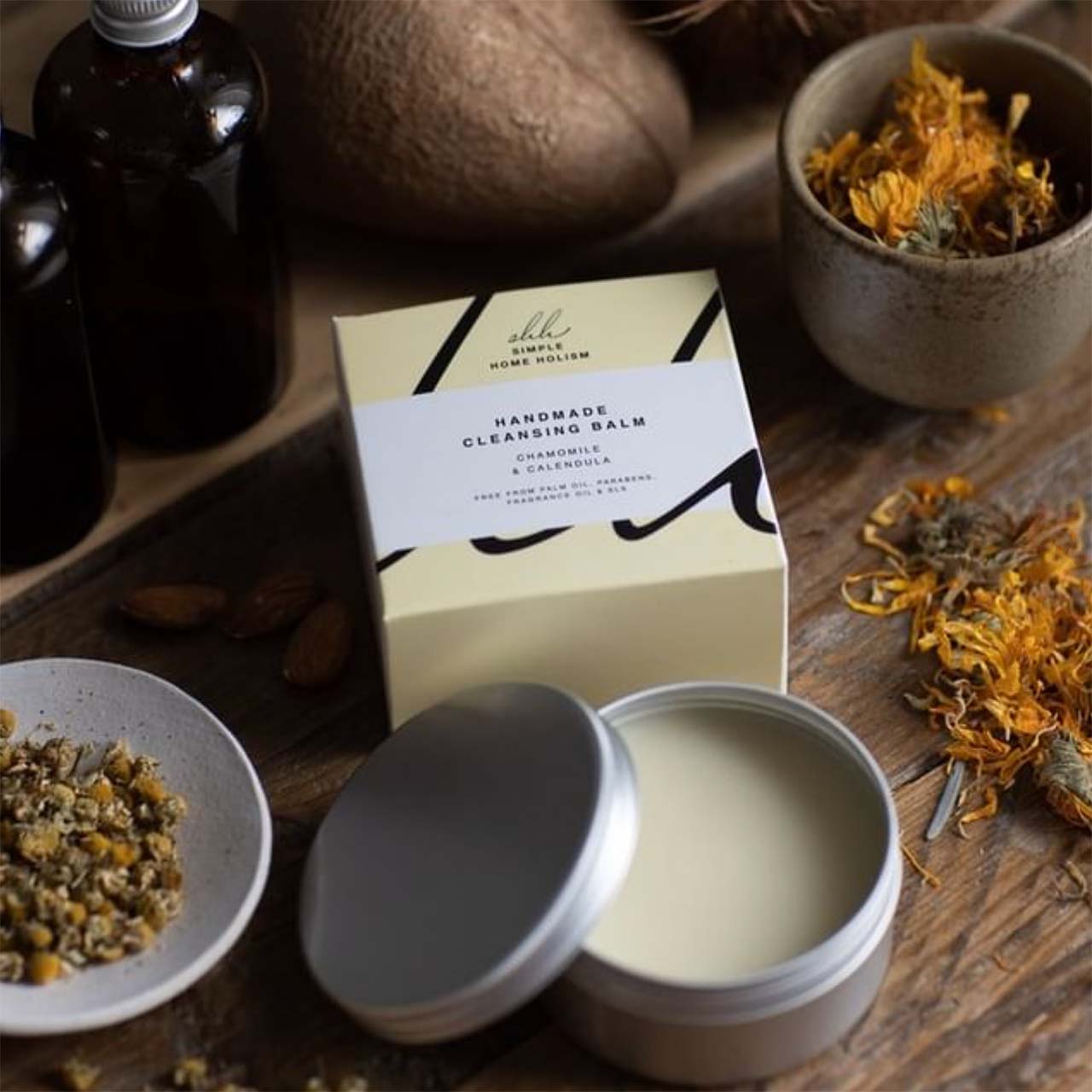 an opened tin of hand balm from simple home holism on a wooden table with natural ingredients and a packaging next to it