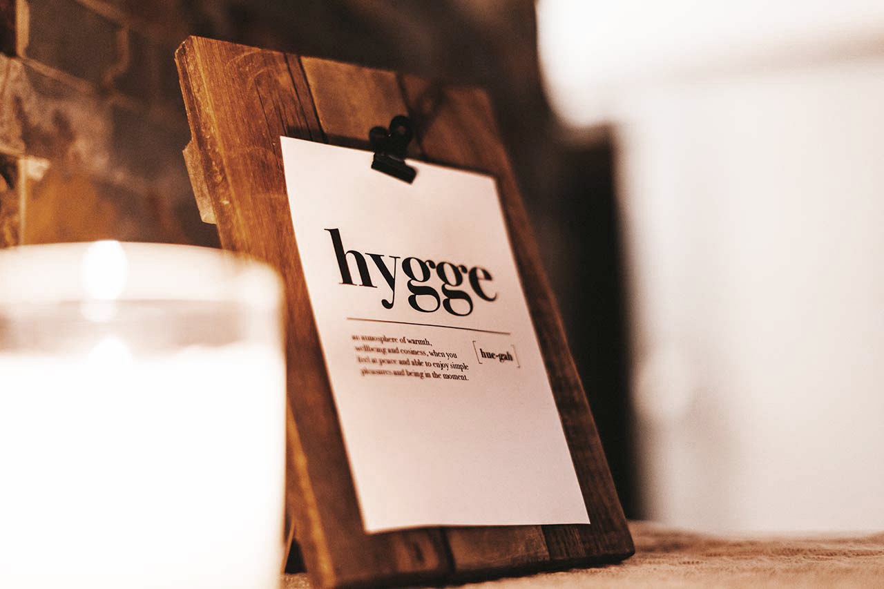 A candle shining on a paper wall hanger with the word hygge and the meaning of it beneath it