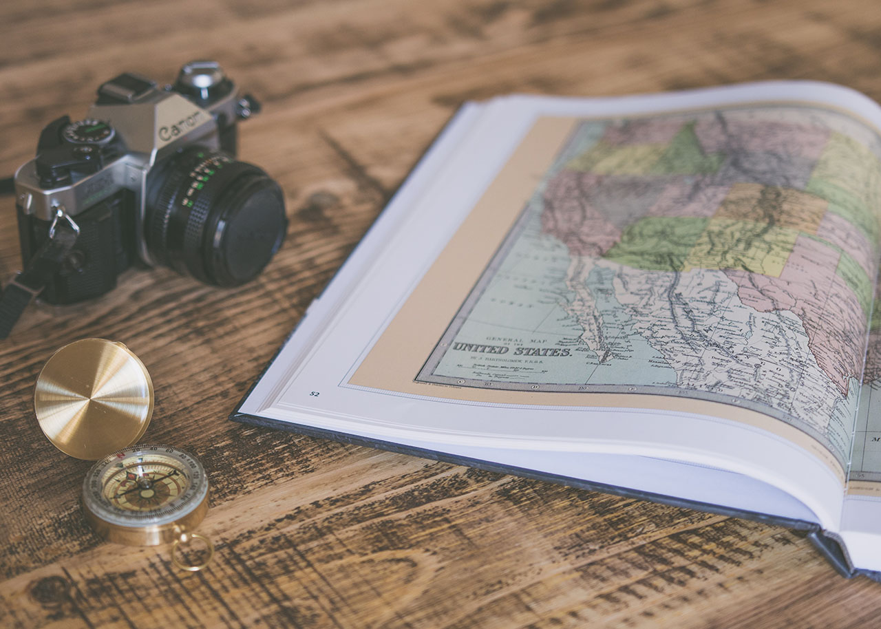 a camera, a compass and a book with maps of the world on a wooden table - slow travel gifts