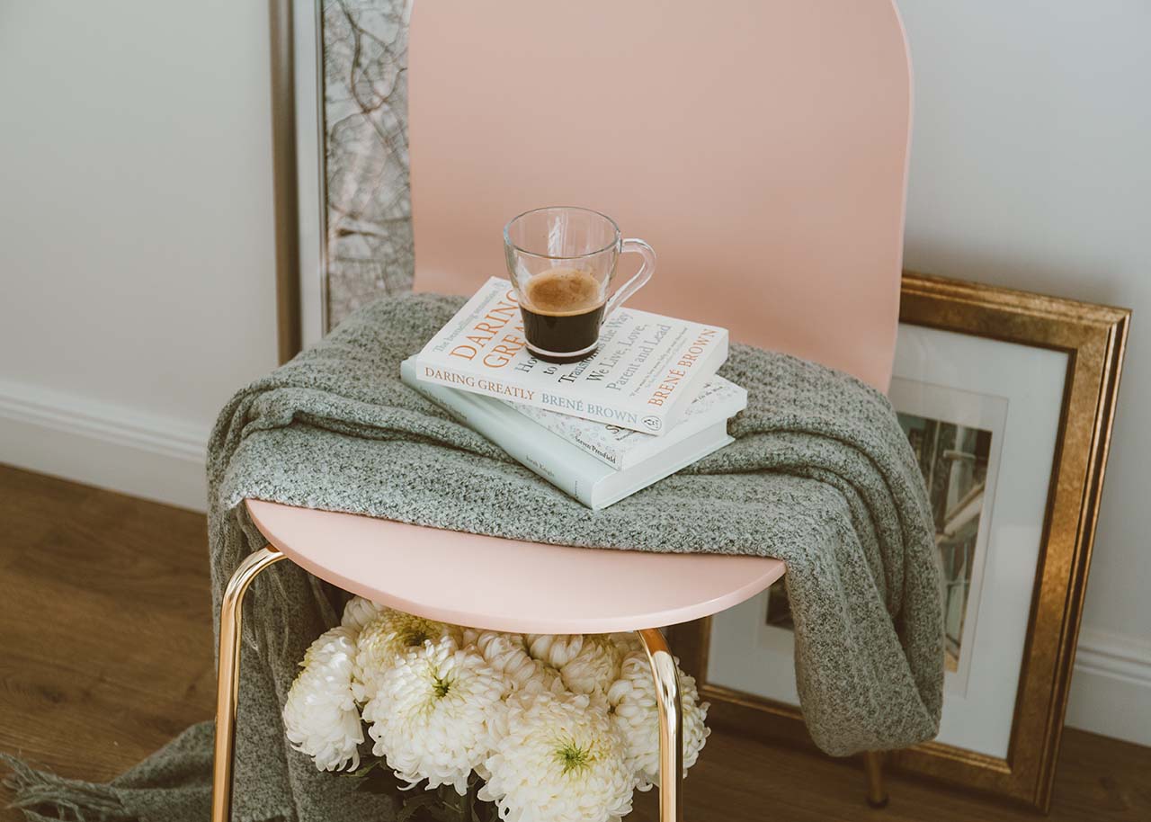 A soft pink chair with white flowers underneath and a grey plaid, a stack of books and a cup of coffee on top - spring interior