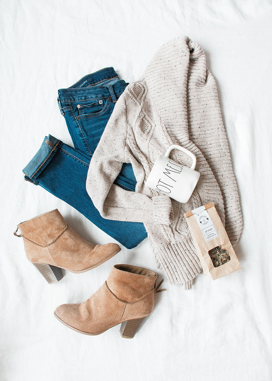 flatlay of timeless wardrobe items: jeans, woolen vest, ankle boots and a tea mug and a package of loose leaf tea