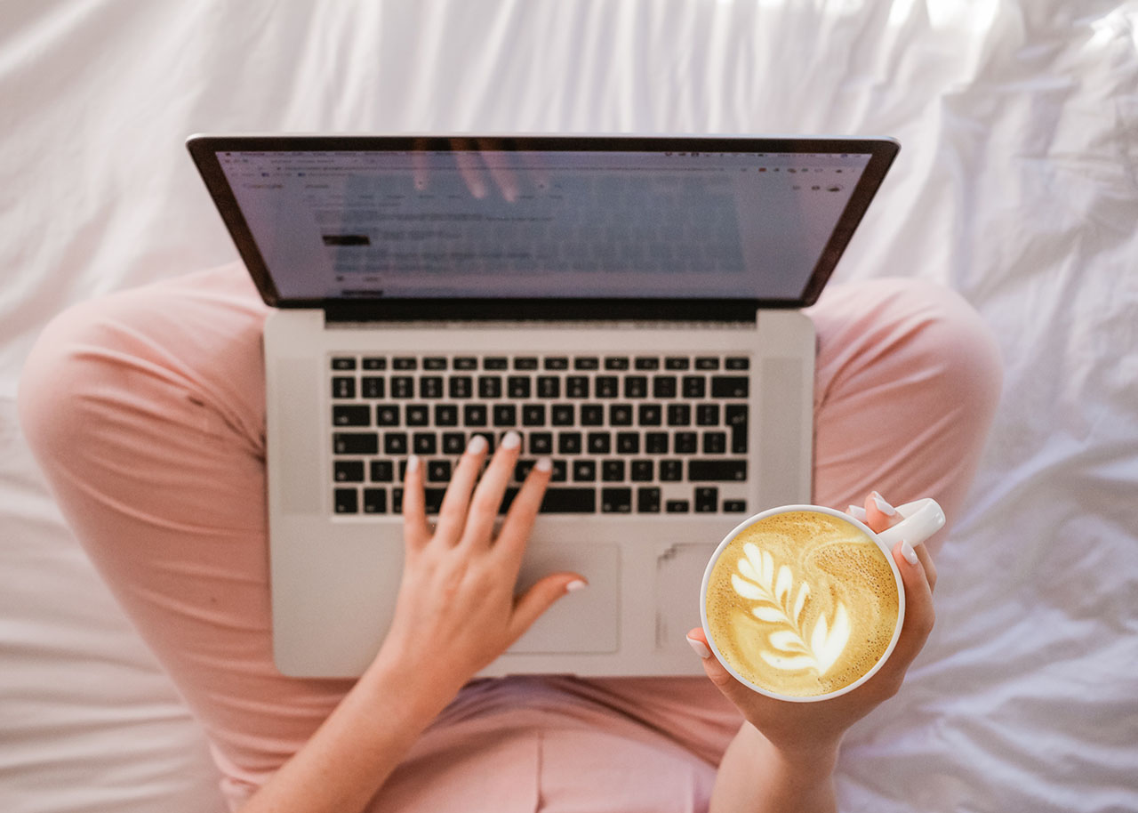a woman sitting on a bed with a laptop on her lap and holding a cup of coffee - hygge at work