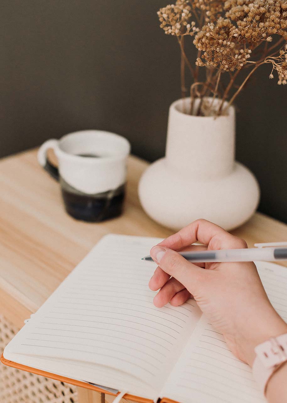 a woman holding a pen to write in an empty notebook, a coffee cup and a vase with a dried flower