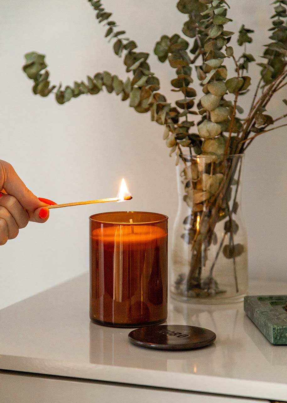a woman's hand lighting a candle next to a vase with eucalyptus - bringing calm into every room