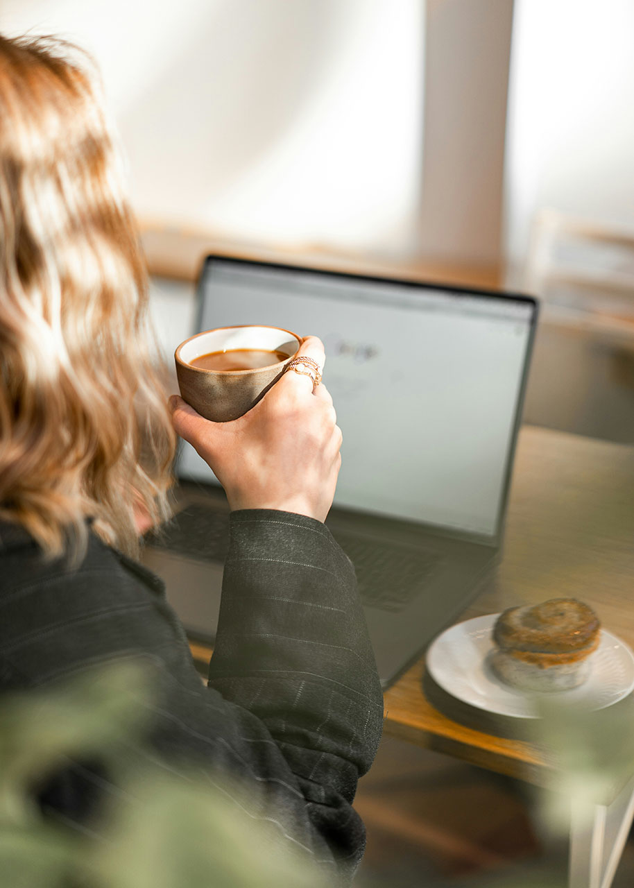 a woman sitting behind a laptop reading while holding a cup of coffee