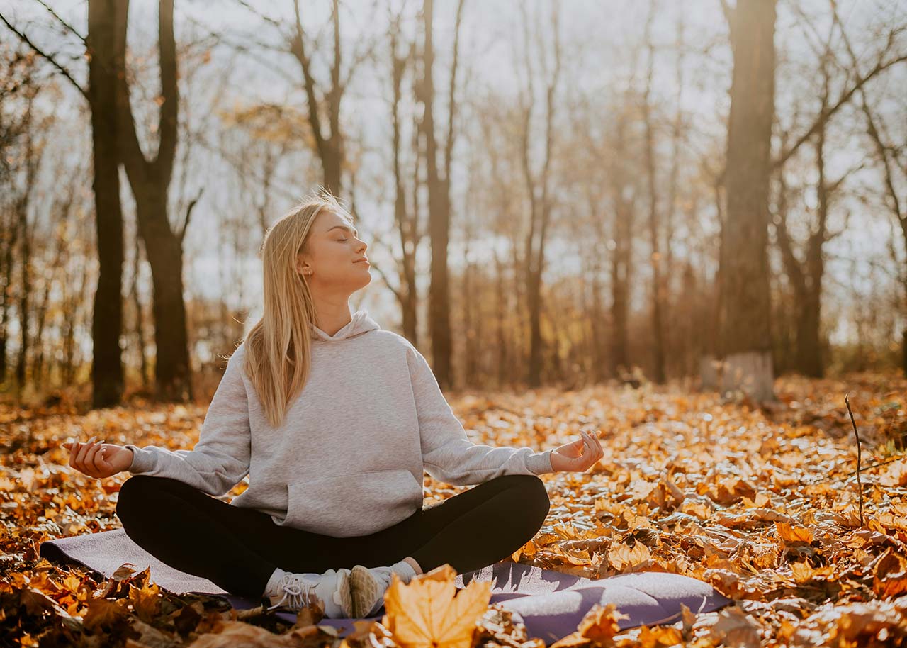 woman sitting in meditation pose in the woods with brown leaves - travel burn out - mindfulness