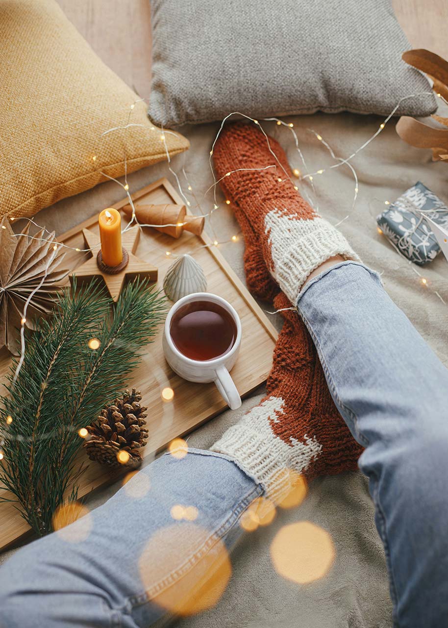 cosy christmas scene with coffee, candle, person wearing woolen socks, fairy lights - holiday gatherings